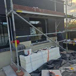 Przemek Najlepszy - Dwóch pracowników na rusztowaniu przy czarnej, ceglanej elewacji budynku. Jeden z nich szpachluje okolice okna, drugi opiera się o rusztowanie. Na ziemi stos białych płyt izolacyjnych i narzędzia.