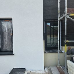 Przemek Najlepszy - Fragment elewacji budynku w trakcie prac wykończeniowych: biały tynk obok ciemnej cegły, okna zabezpieczone folią, rusztowanie i pracownik przy dolnej części ściany.