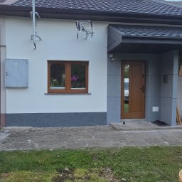 Przemek Najlepszy - Wykończona elewacja domu jednorodzinnego z szarą podmurówką i białą górną częścią, brązowe okno z kwiatami, szare wejście z brązowymi drzwiami, antena na ścianie, trawnik z roślinami.