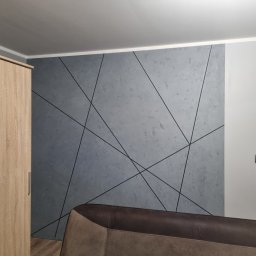 Przemek Najlepszy - Szara ściana z geometrycznym wzorem z czarnych linii, widoczny fragment szafy z jasnego drewna i brązowy narożnik sofy. W tle widoczne wnętrze kuchni.