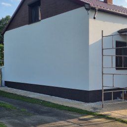 Przemek Najlepszy - Świeżo otynkowana ściana boczna domu jednorodzinnego z brązowym cokołem, widoczne rusztowanie, okno zabezpieczone folią.