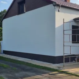 Świeżo otynkowana ściana boczna domu jednorodzinnego z brązowym cokołem, widoczne rusztowanie, okno zabezpieczone folią.