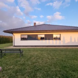 Jednopiętrowy dom z jasną elewacją i ciemnymi obramowaniami okien, widoczny z perspektywy trawnika z ławką, w tle rząd drzew i błękitne niebo z chmurami, oświetlony popołudniowym słońcem.