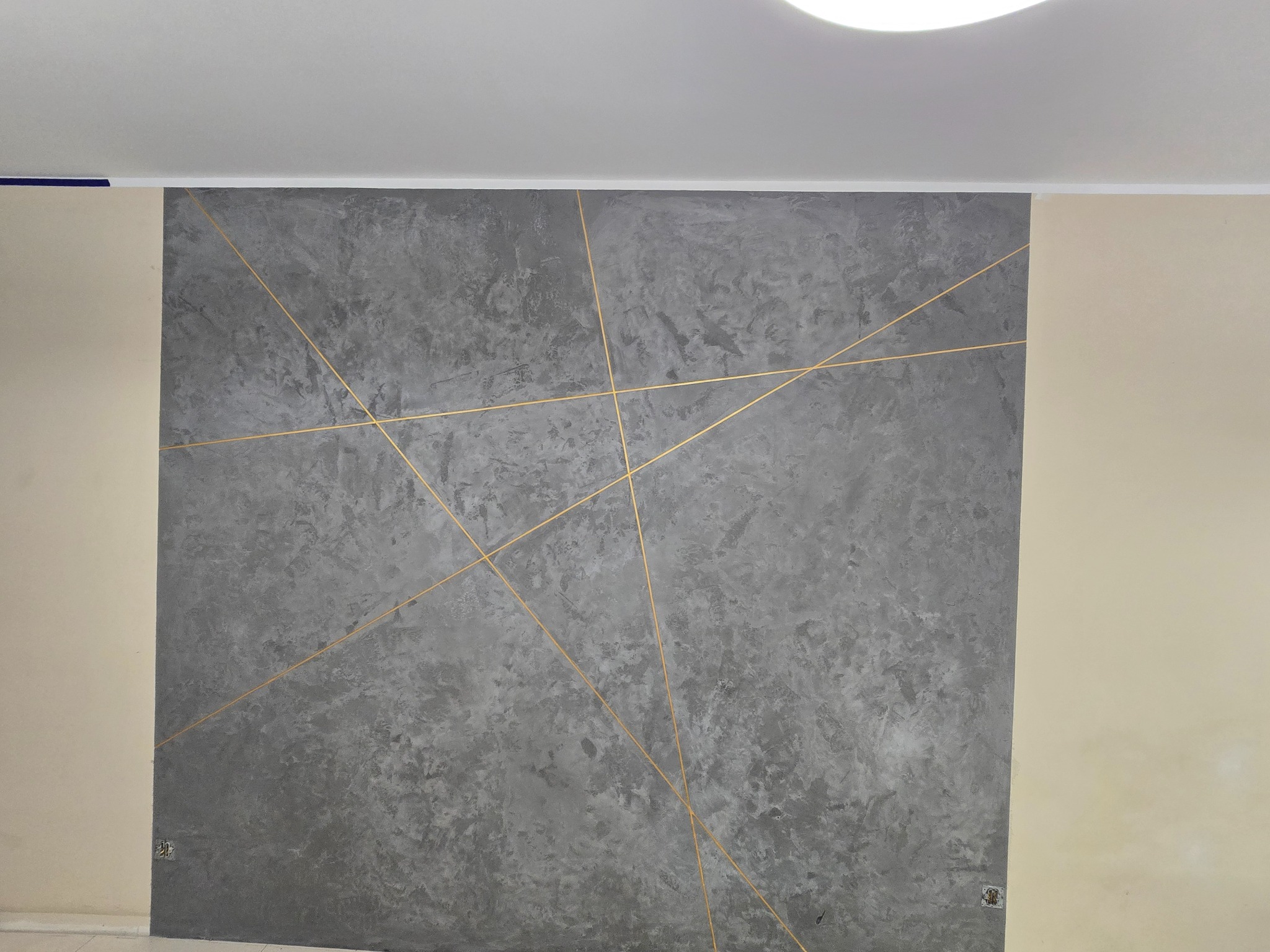 Szara ściana z dekoracyjną fakturą i złotymi liniami tworzącymi geometryczny wzór, widoczny fragment sufitu i sąsiedniej ściany w kolorze ecru.