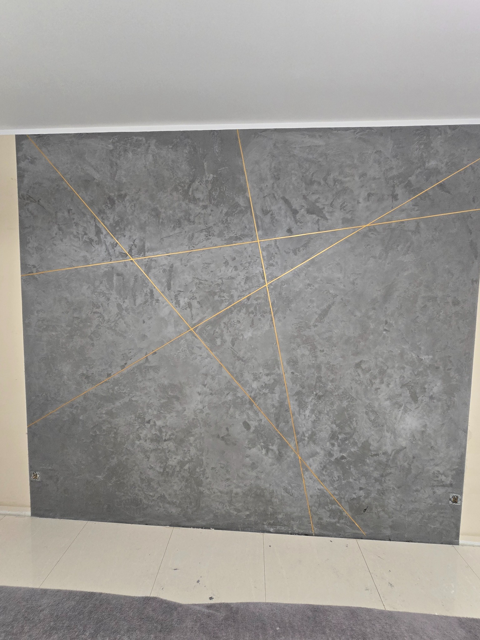 Szara ściana z dekoracyjną fakturą i złotymi liniami tworzącymi geometryczny wzór, widoczne gniazdka elektryczne na ścianie, jasna podłoga i fragment szarego dywanu.