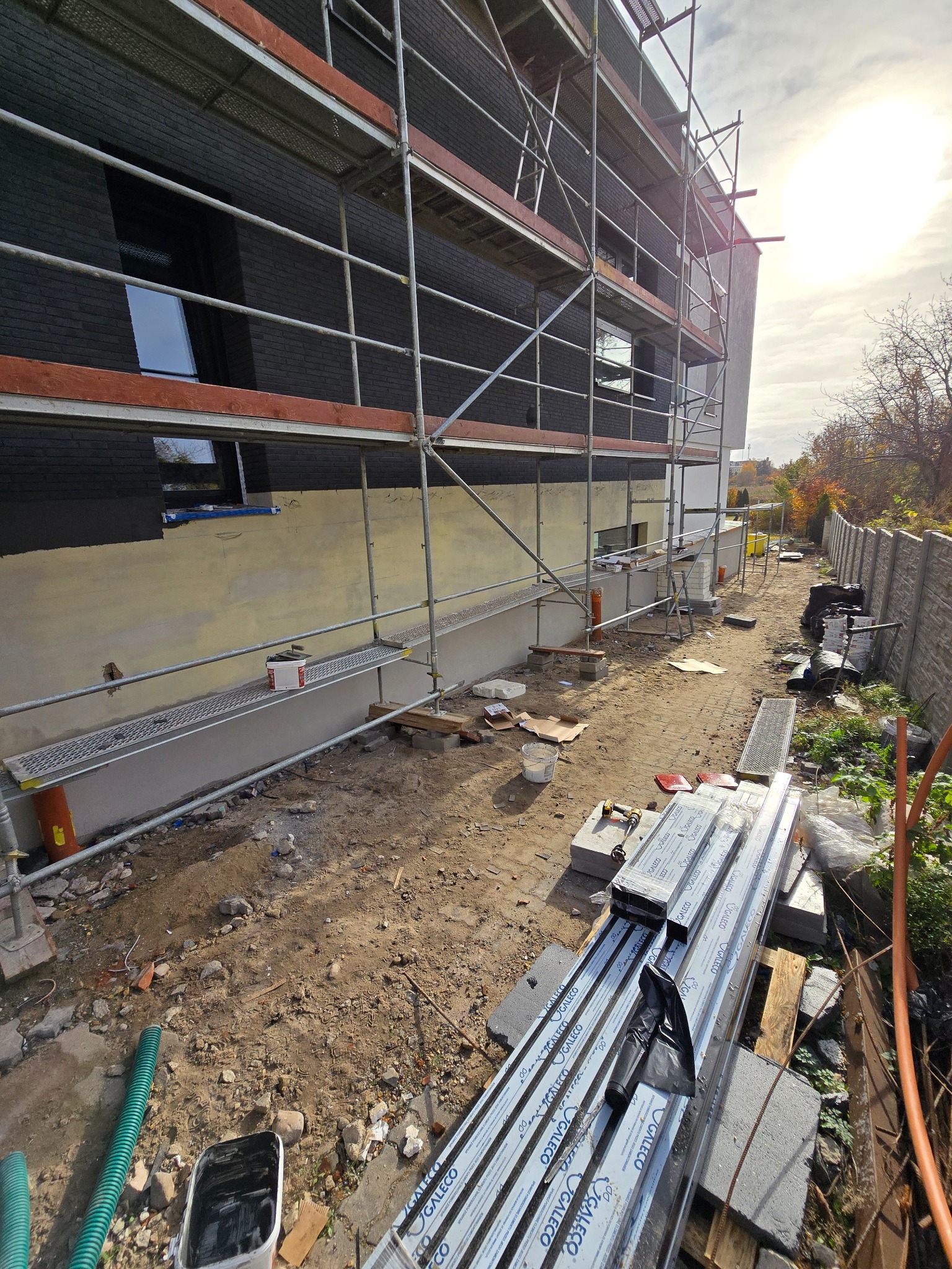 Budynek w trakcie prac elewacyjnych, widoczne rusztowania, częściowo pomalowana ściana na ciemny kolor, przygotowane profile aluminiowe GALECO, teren budowy z narzędziami i materiałami.