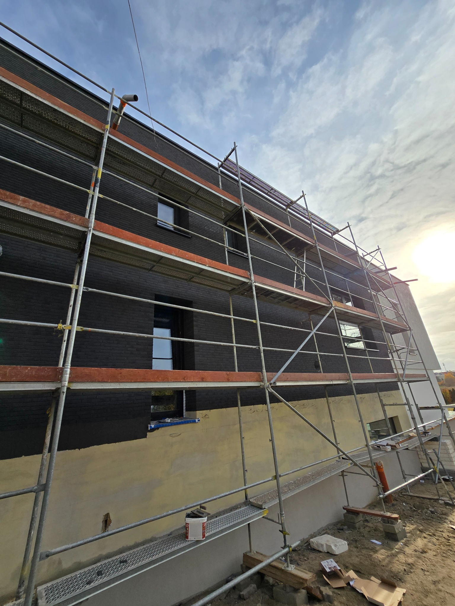 Elewacja domu w trakcie prac wykończeniowych z czarną cegłą klinkierową, widoczne rusztowania, fragment ściany z nałożonym tynkiem podkładowym, błękitne niebo z chmurami w tle.