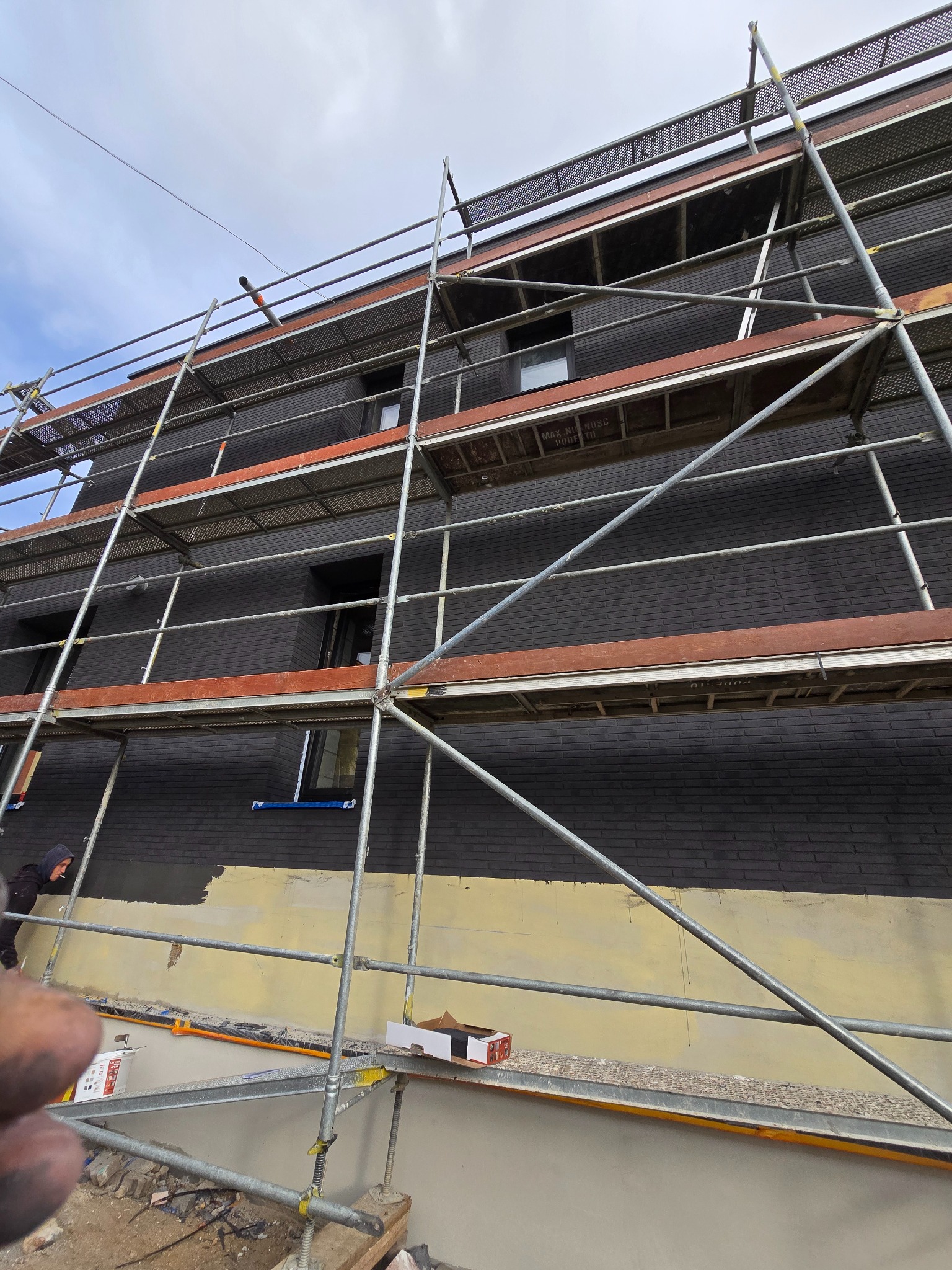 Czarny budynek z cegły w trakcie prac elewacyjnych, widoczne rusztowania, dolna część ściany pokryta warstwą jasnego tynku, pracownik w kapturze z papierosem w ustach.