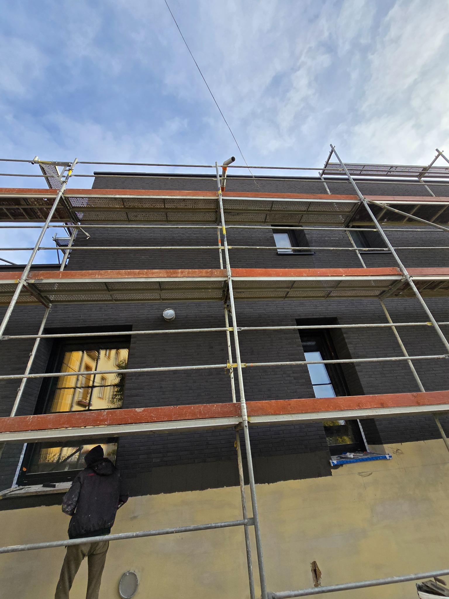 Pracownik w czarnej kurtce na rusztowaniu maluje elewację budynku na ciemny kolor, widoczne okna i niebo z chmurami.