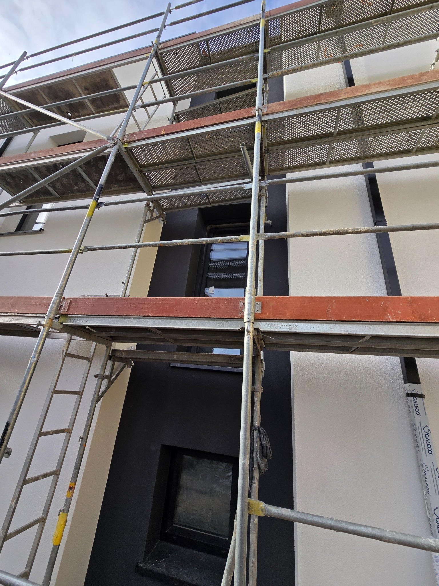 Elewacja budynku w trakcie prac wykończeniowych, widoczne rusztowanie, fragmenty tynku o różnej fakturze i kolorystyce, okna z czarnymi ramami.