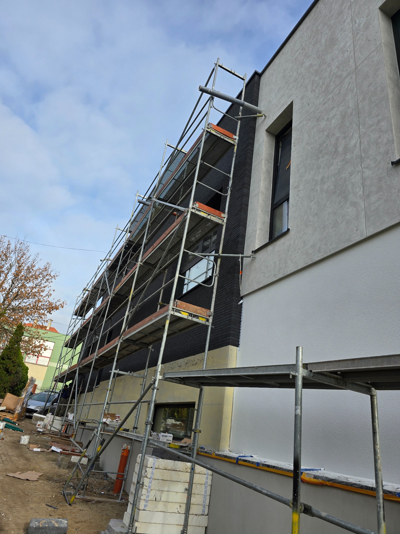 Dwukolorowa elewacja budynku w trakcie prac wykończeniowych, z widocznym rusztowaniem i materiałami budowlanymi na ziemi.