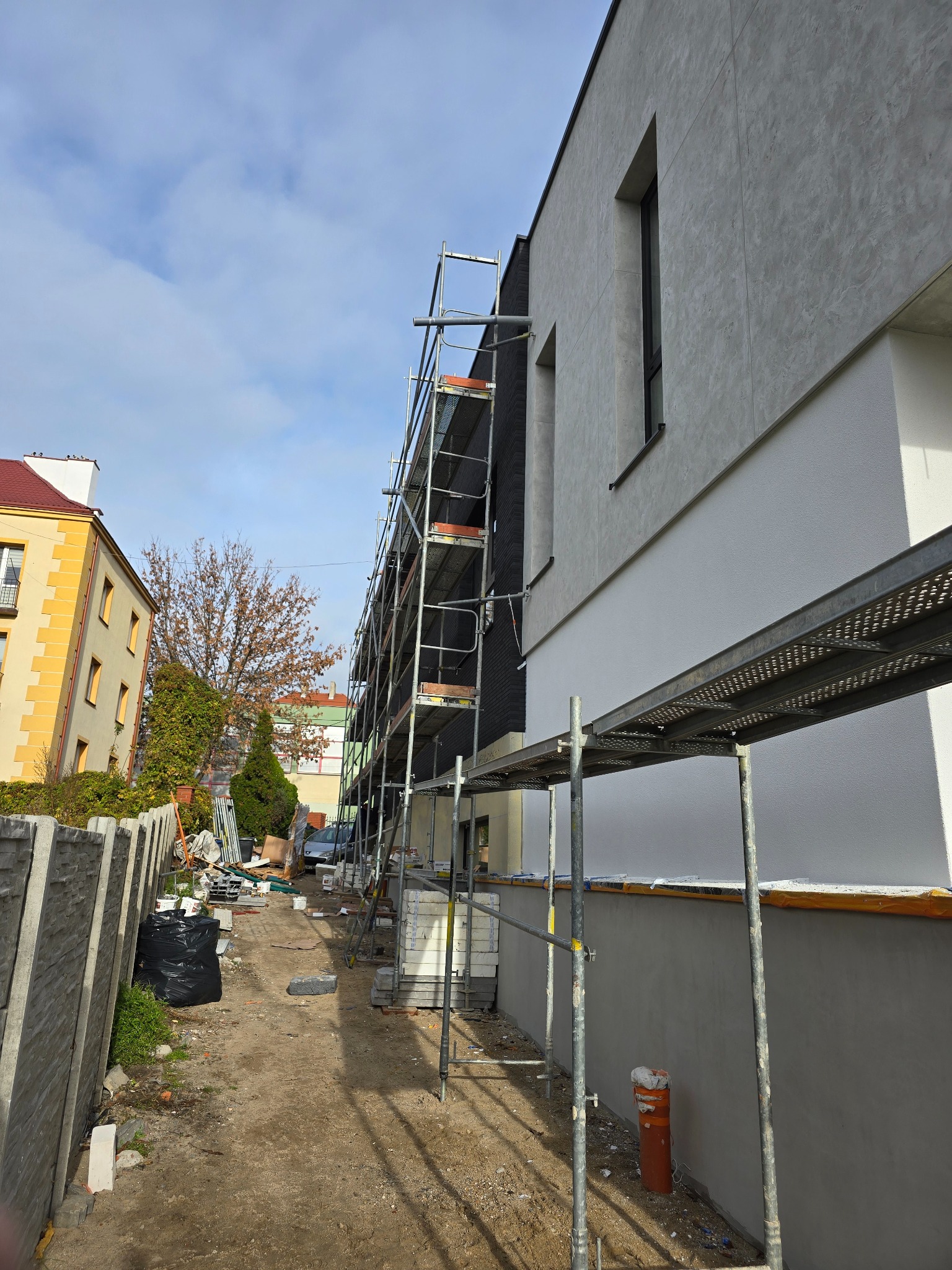 Budynek w trakcie prac wykończeniowych elewacji, widoczne rusztowania, fragmenty otynkowanej ściany z oknami, przygotowany teren wokół budynku.