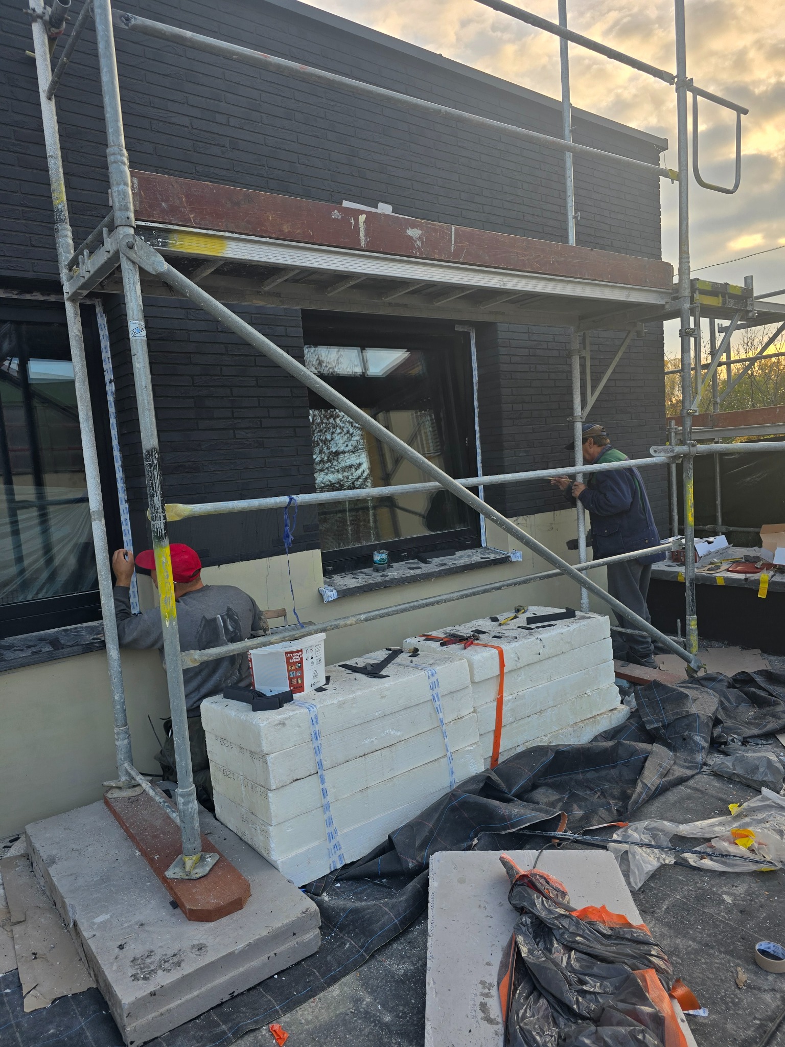 Dwóch pracowników na rusztowaniu przy czarnej, ceglanej elewacji budynku. Jeden z nich szpachluje okolice okna, drugi opiera się o rusztowanie. Na ziemi stos białych płyt izolacyjnych i narzędzia.