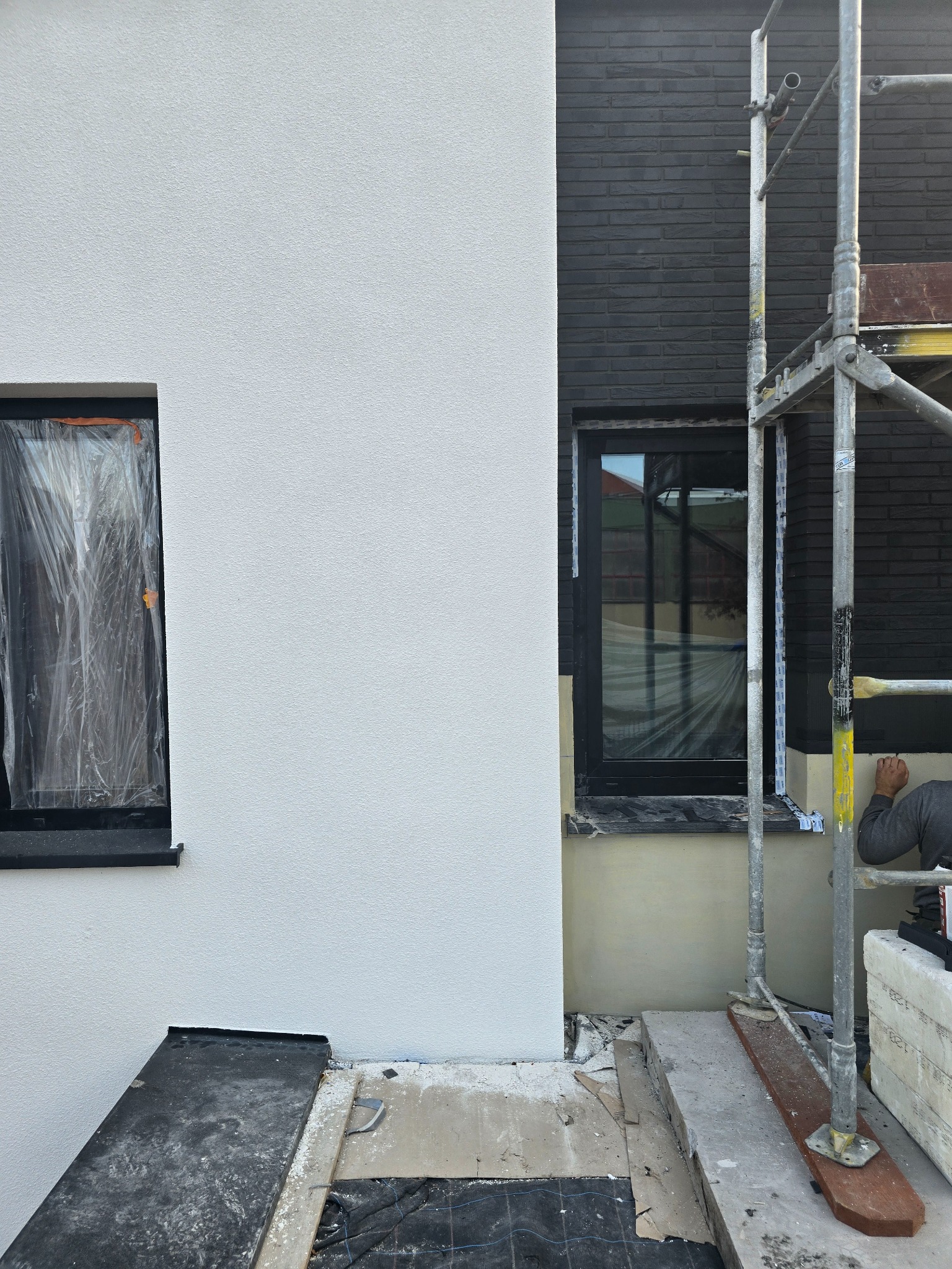 Fragment elewacji budynku w trakcie prac wykończeniowych: biały tynk obok ciemnej cegły, okna zabezpieczone folią, rusztowanie i pracownik przy dolnej części ściany.