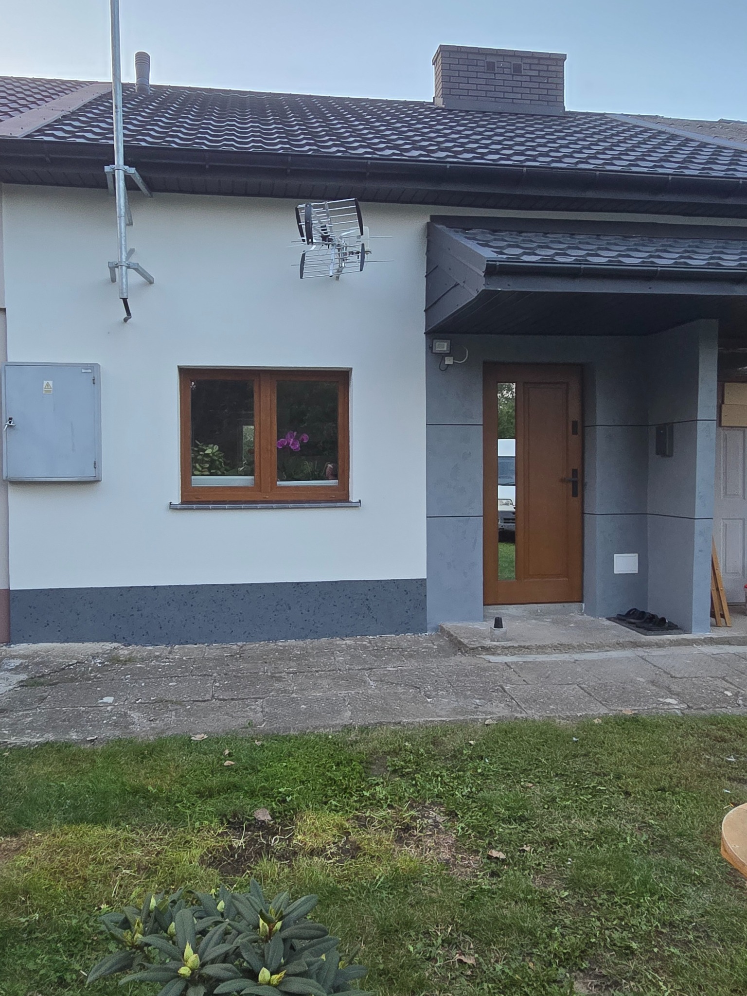 Dom jednorodzinny po remoncie elewacji, z widoczną anteną telewizyjną, oknem z drewnianą ramą i roślinami wewnątrz, oraz wejściem z szarą okładziną i brązowymi drzwiami, z elementami wykończenia...