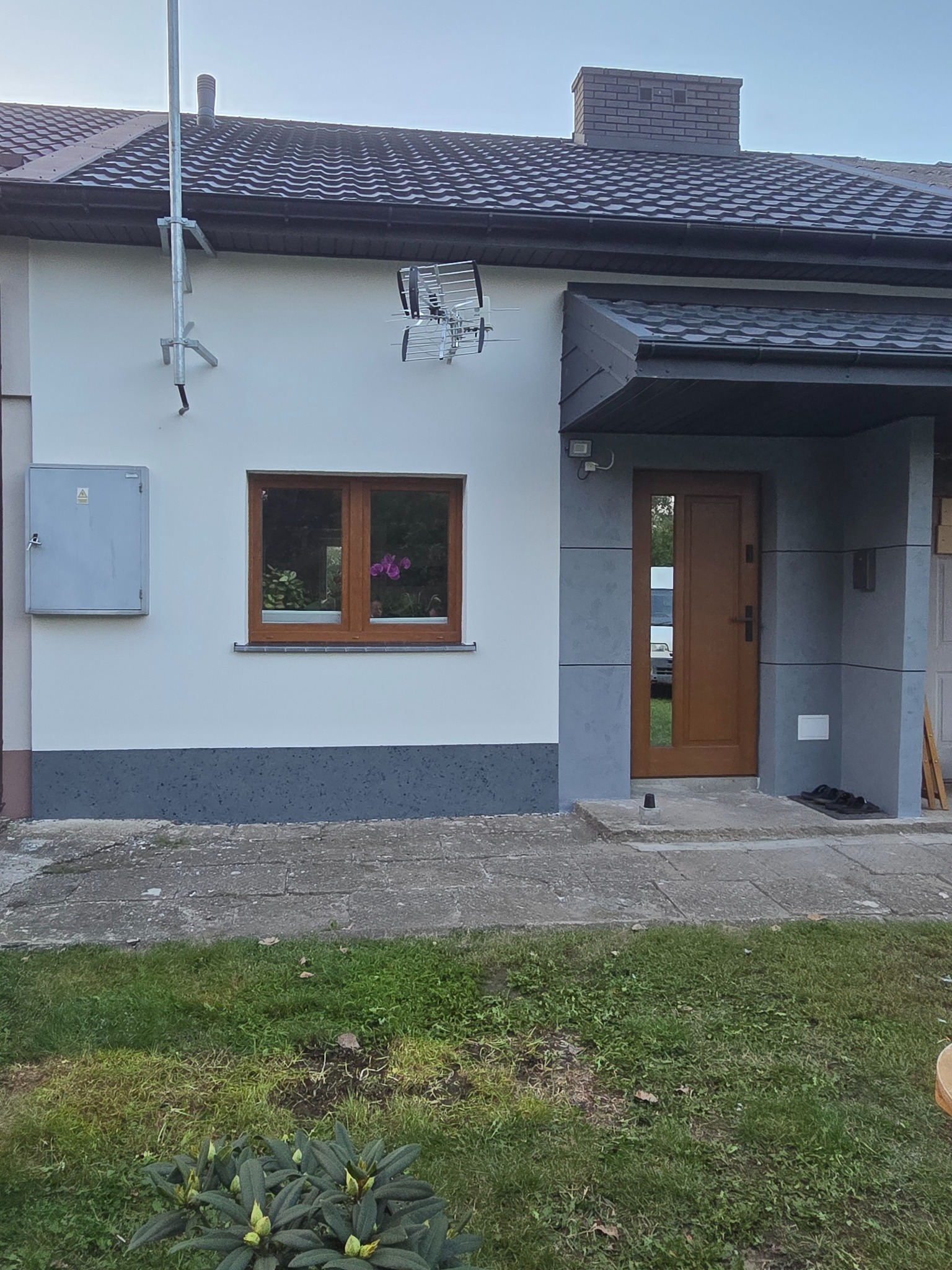 Wykończona elewacja domu jednorodzinnego z szarą podmurówką i białą górną częścią, brązowe okno z kwiatami, szare wejście z brązowymi drzwiami, antena na ścianie, trawnik z roślinami.