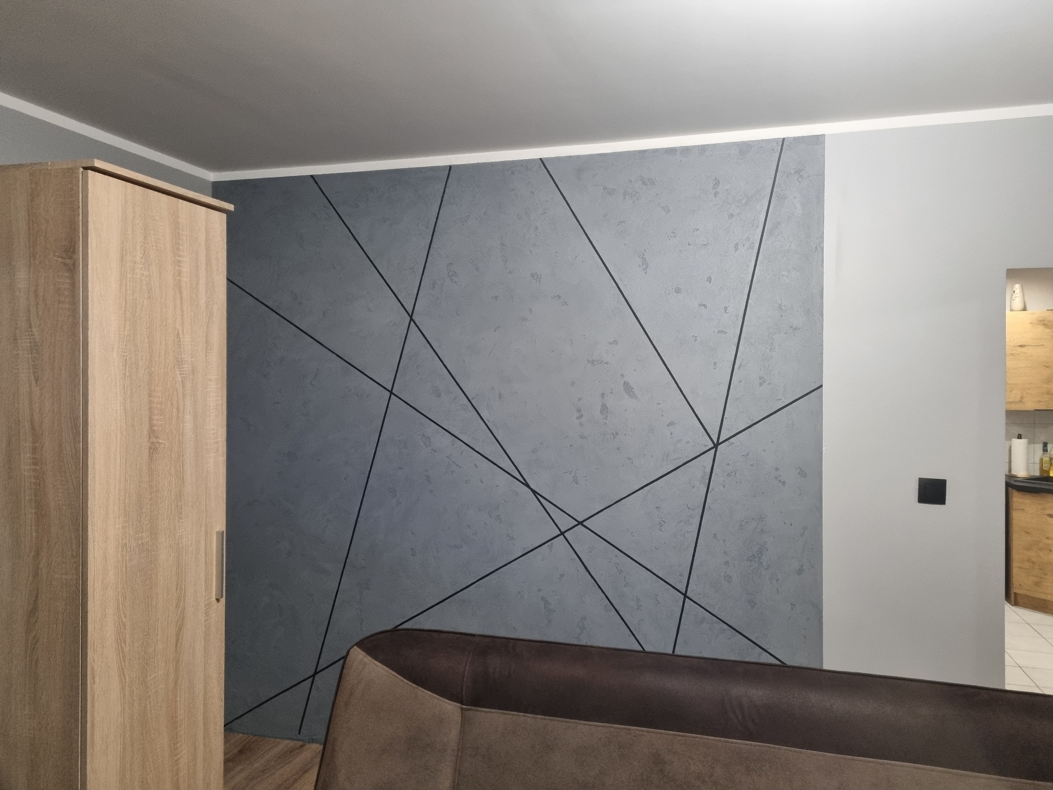 Szara ściana z geometrycznym wzorem z czarnych linii, widoczny fragment szafy z jasnego drewna i brązowy narożnik sofy. W tle widoczne wnętrze kuchni.