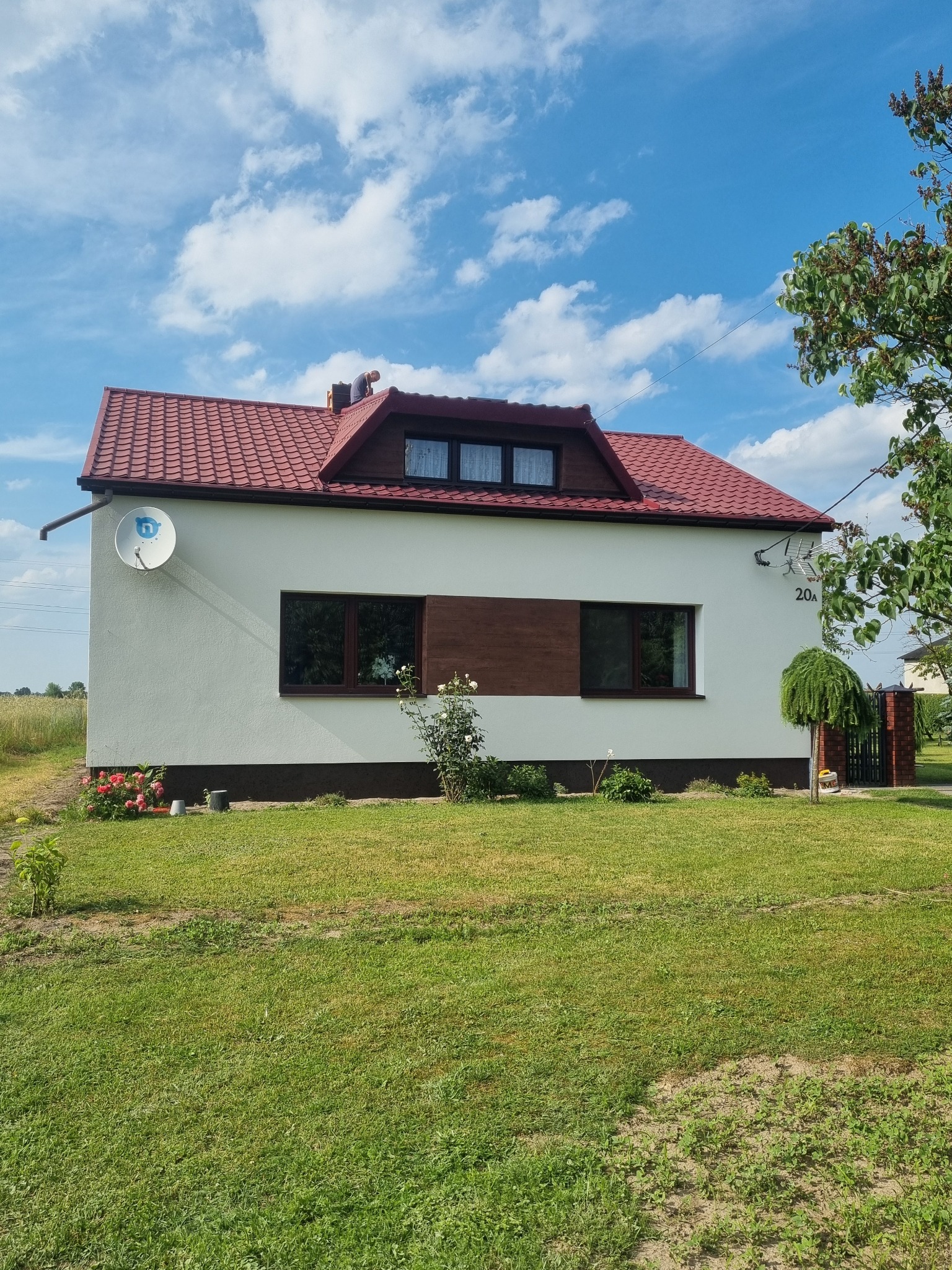 Biały dom jednorodzinny z czerwoną dachówką, brązowymi oknami i elewacją po renowacji, na zadbanym trawniku w słoneczny dzień, z widocznym numerem domu '20a'.