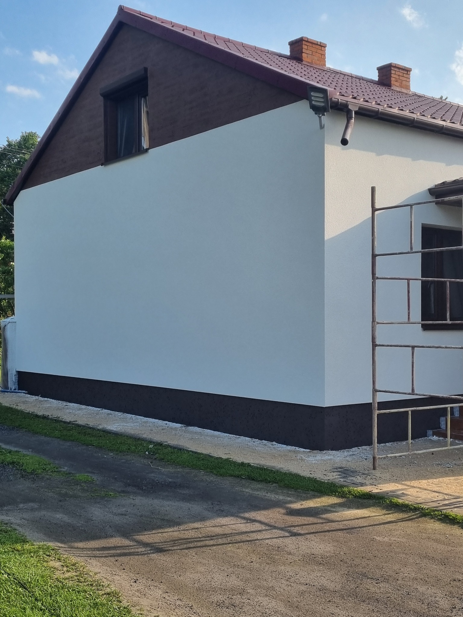 Świeżo otynkowana ściana boczna domu jednorodzinnego z brązowym cokołem, widoczne rusztowanie, okno zabezpieczone folią.