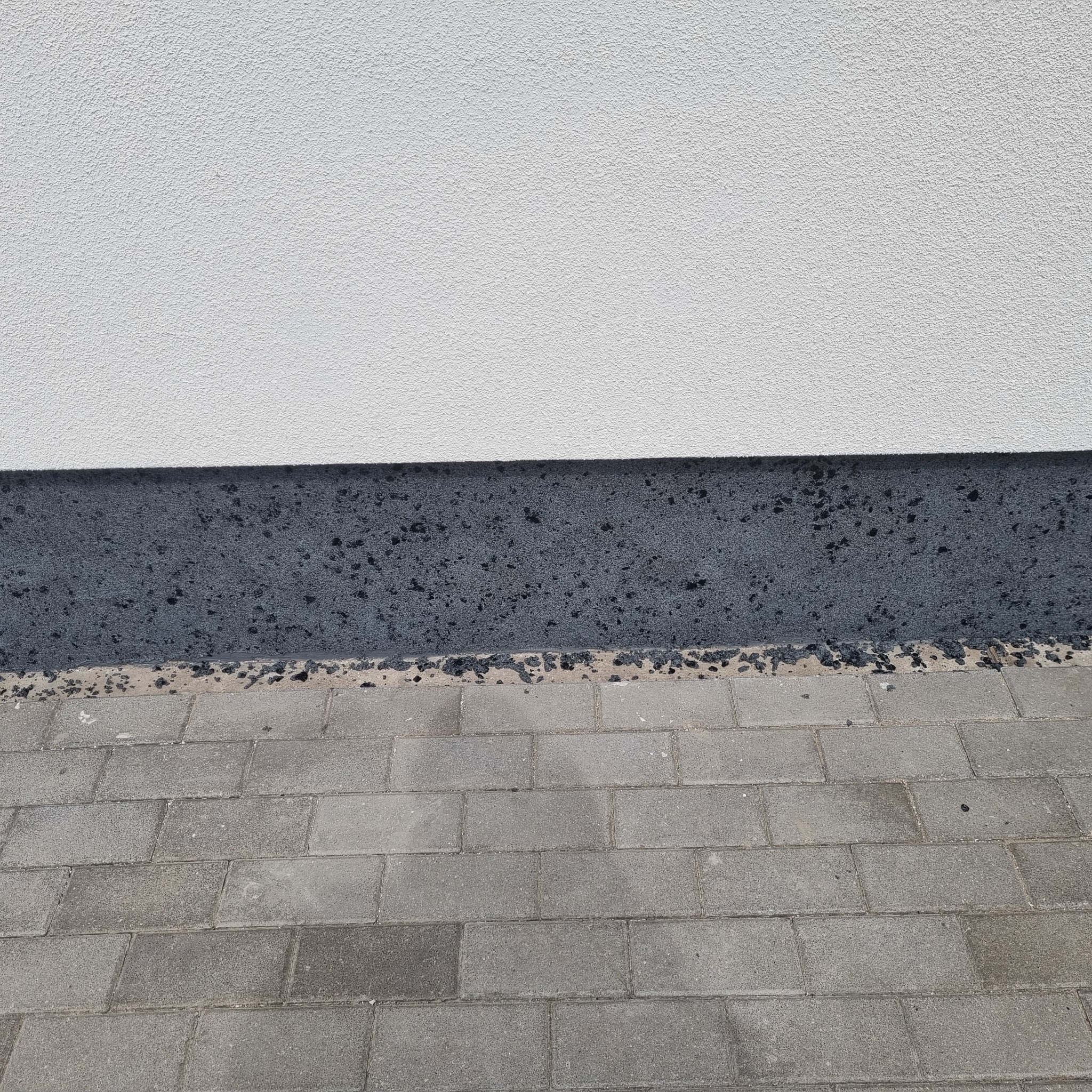 Szara elewacja wykończona tynkiem strukturalnym, z ciemnoszarym cokołem z widocznymi elementami kruszywa, obok ułożona szara kostka brukowa.