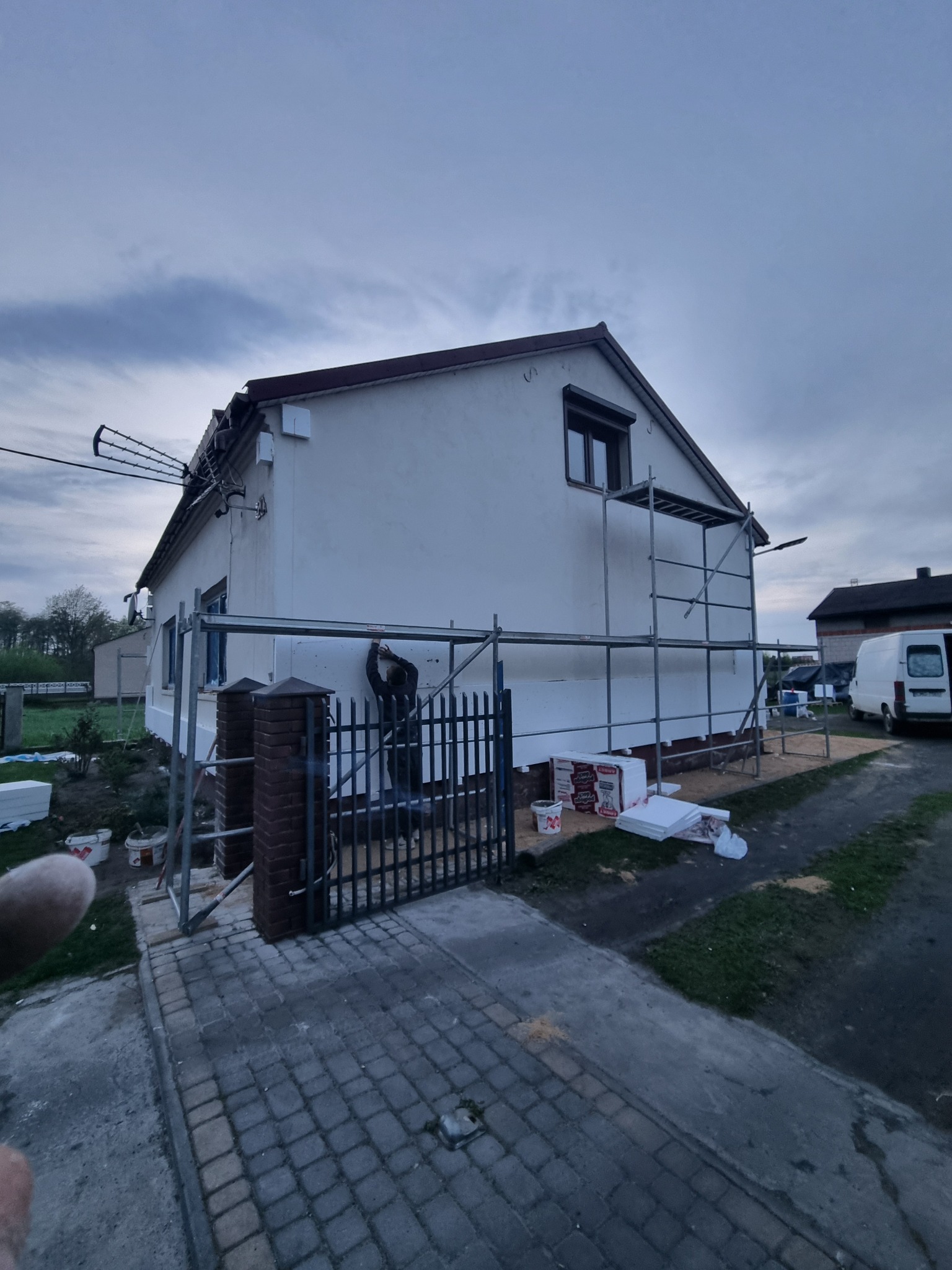 Ocieplanie elewacji domu jednorodzinnego styropianem, widoczne rusztowanie, materiały budowlane i pracownik w trakcie montażu, pochmurne niebo.