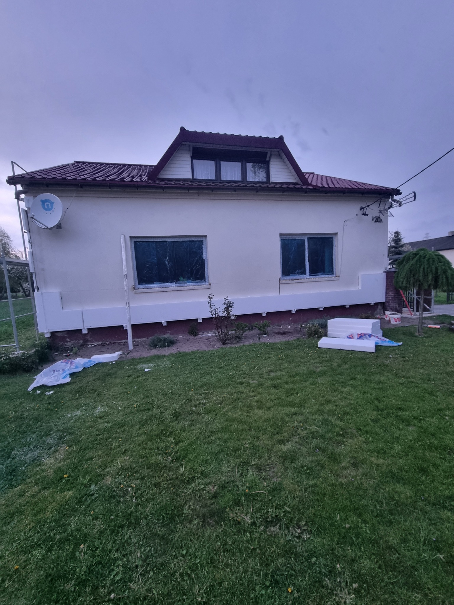 Dom jednorodzinny w trakcie prac przygotowawczych do ocieplenia elewacji, widoczne zabezpieczone folią okna, częściowo zamontowane płyty izolacyjne przy fundamentach oraz stos styropianu na trawniku.