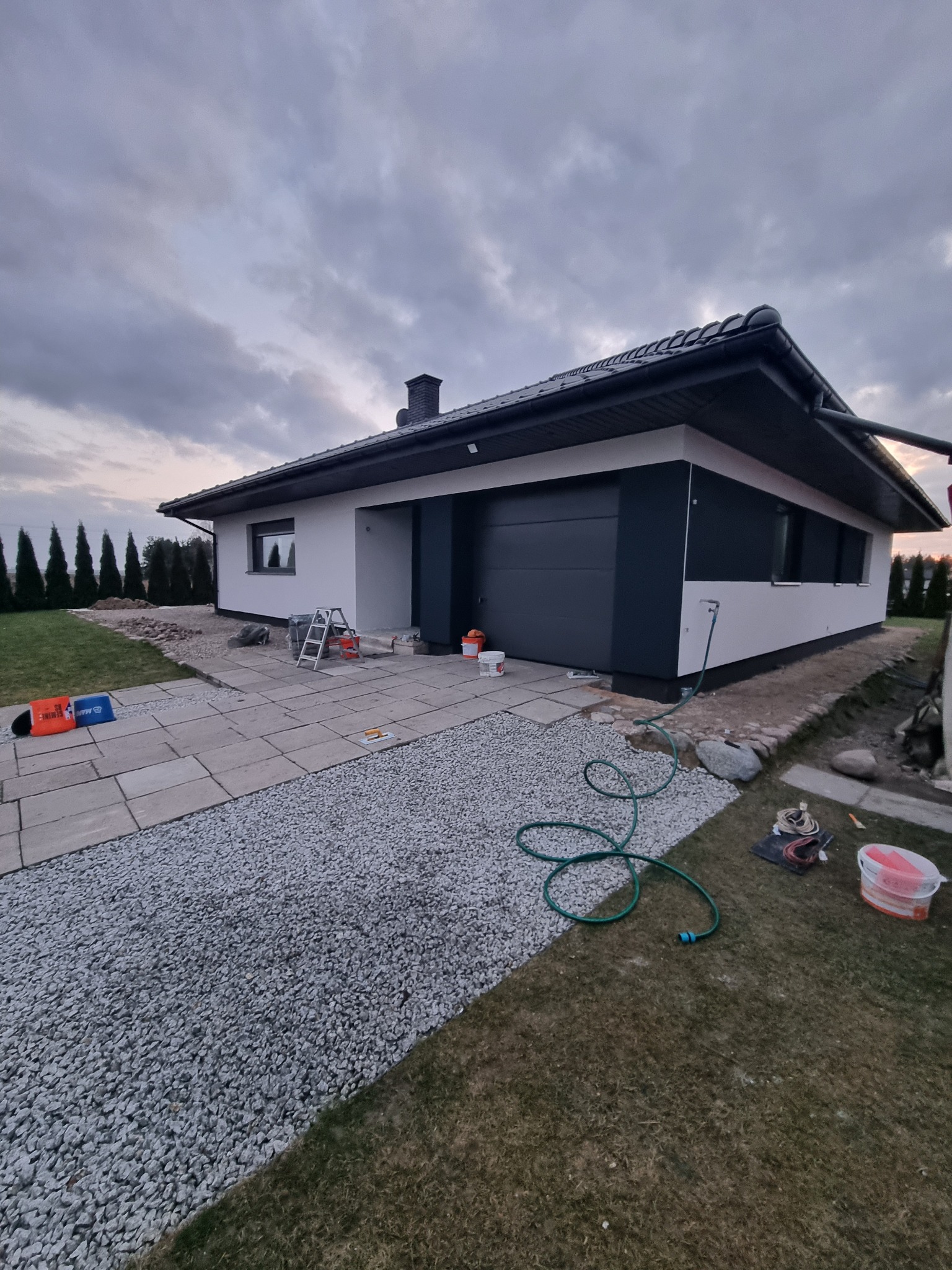 Jednopiętrowy dom z garażem w trakcie prac wykończeniowych elewacji, widoczne narzędzia, wiaderka z farbą, drabina, kostka brukowa i zielony wąż ogrodowy na trawniku.