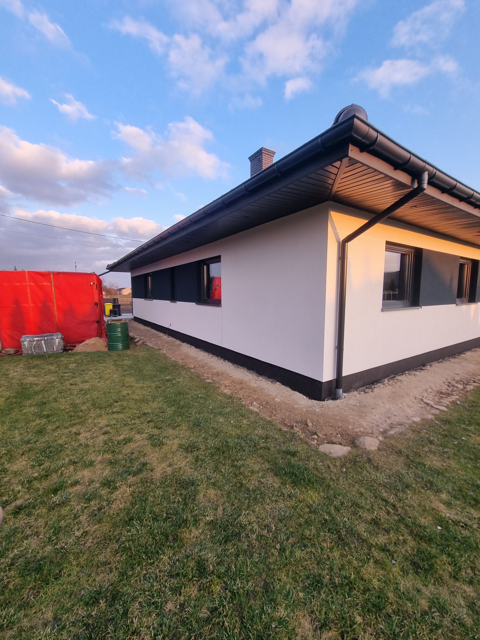 Świeżo wykończony, parterowy dom z białą elewacją i czarnymi detalami, w trakcie prac wykończeniowych wokół budynku, na trawniku widoczne beczki i zabezpieczenia budowlane.