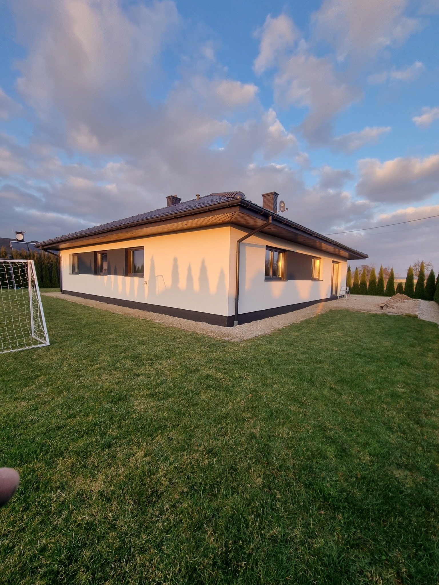 Nowoczesny dom jednorodzinny z białą elewacją i ciemnymi oknami, oświetlony zachodzącym słońcem, z widocznym trawnikiem i siatką do piłki nożnej.