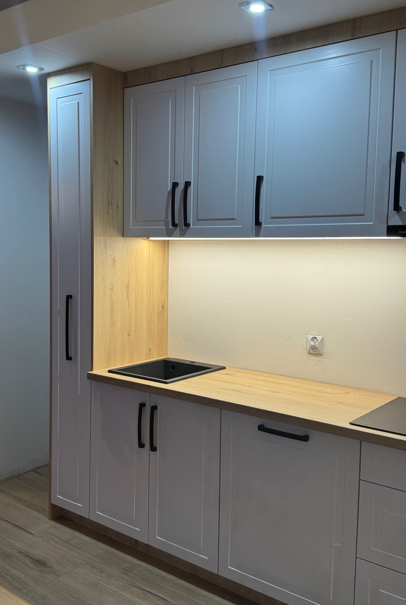 Nowoczesna zabudowa kuchenna z białymi frontami, czarnymi uchwytami i blatem w kolorze jasnego drewna, oświetlenie podszafkowe LED, widoczny zlew i gniazdko elektryczne na ścianie.