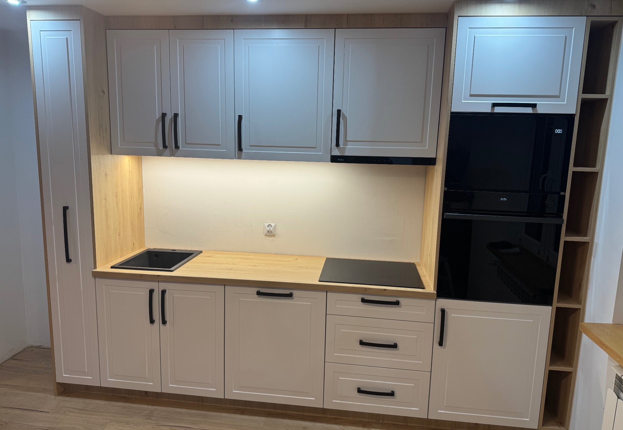 Nowoczesna zabudowa kuchenna z białymi frontami, czarnymi uchwytami i blatem imitującym drewno, z wbudowanym piekarnikiem i płytą grzewczą.