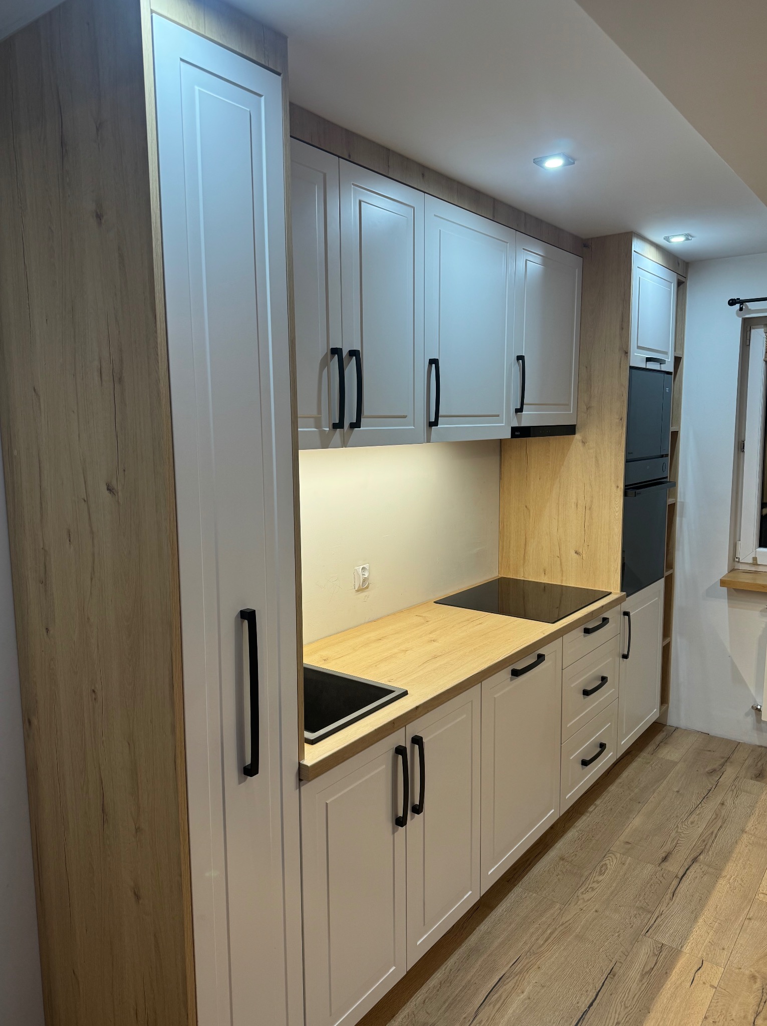 Nowoczesna zabudowa kuchenna z białymi frontami i drewnopodobnym wykończeniem, czarne, minimalistyczne uchwyty, płyta indukcyjna w blacie, oświetlenie podszafkowe, drewniana podłoga.