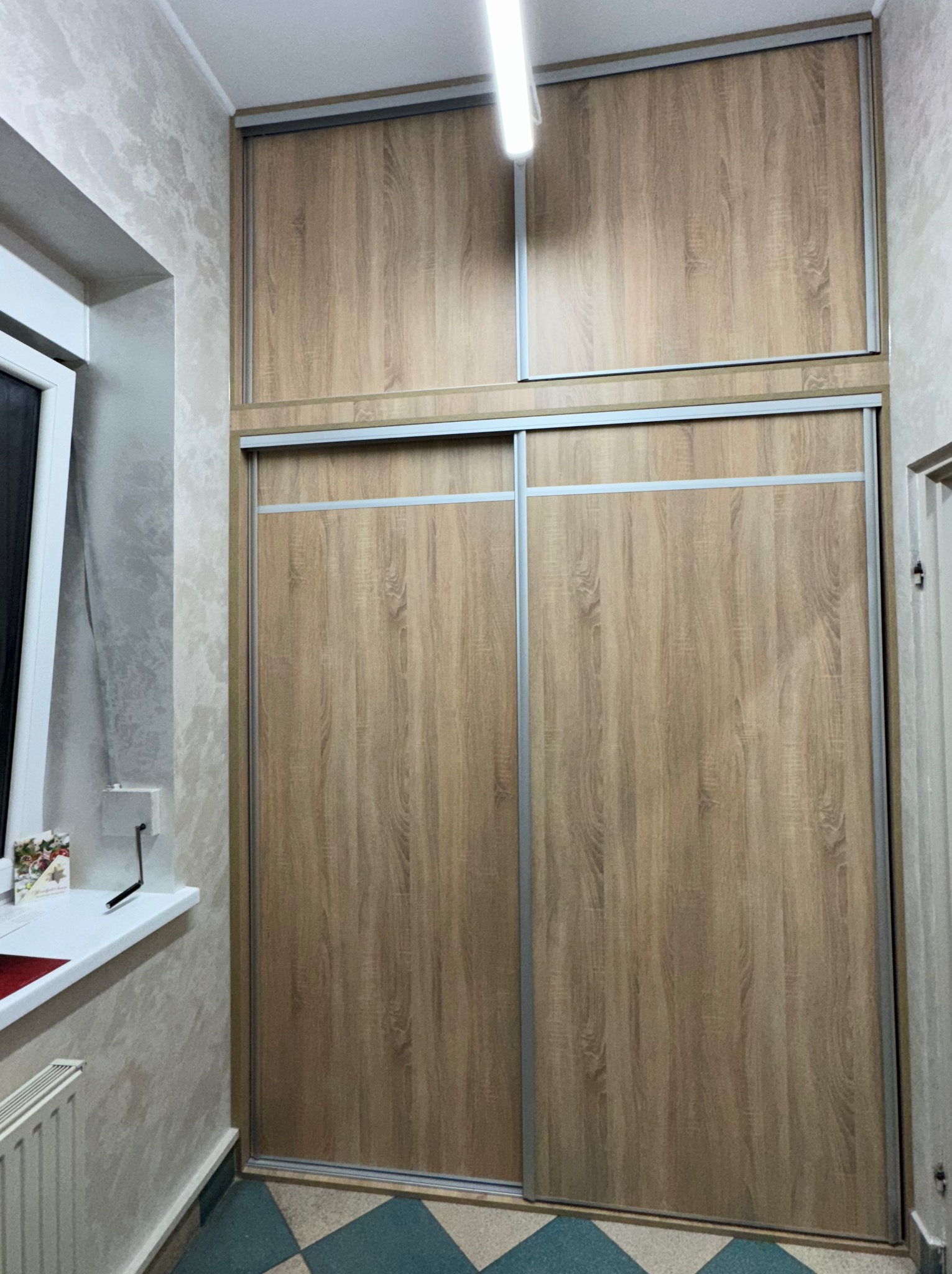 Dwudrzwiowa szafa przesuwna z drewnopodobnymi frontami i nadstawką, widoczna w korytarzu z oknem po lewej stronie i wzorzystą tapetą.