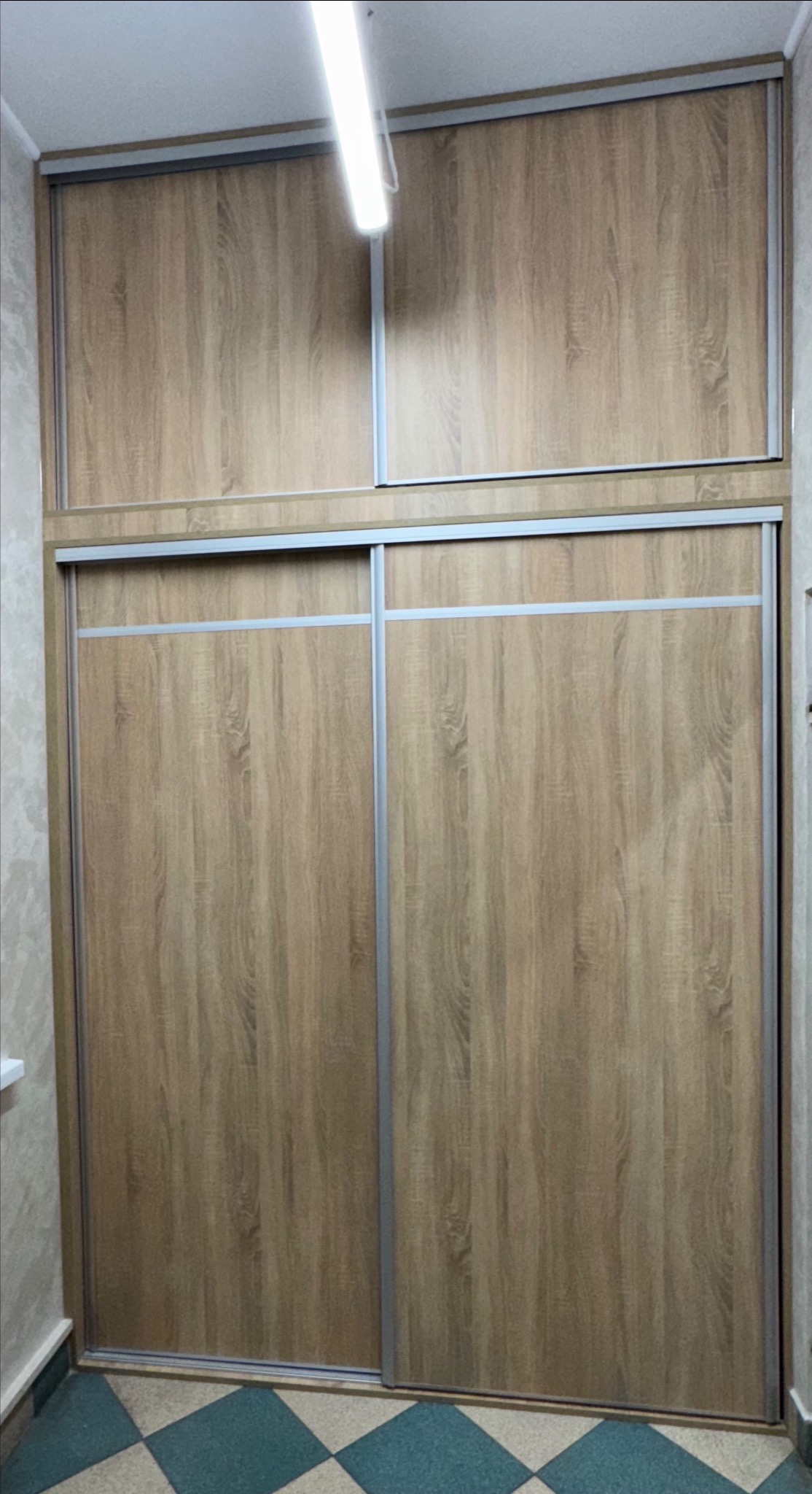 Wysoka, zabudowana szafa przesuwna z drewnopodobnymi frontami i srebrnymi profilami, z dodatkową szafką nad główną częścią, oświetlona górnym światłem.