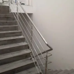 Wewnętrzne schody wyłożone szarymi płytkami imitującymi drewno, zabezpieczone nowoczesną, minimalistyczną poręczą ze stali nierdzewnej z poprzecznymi prętami.