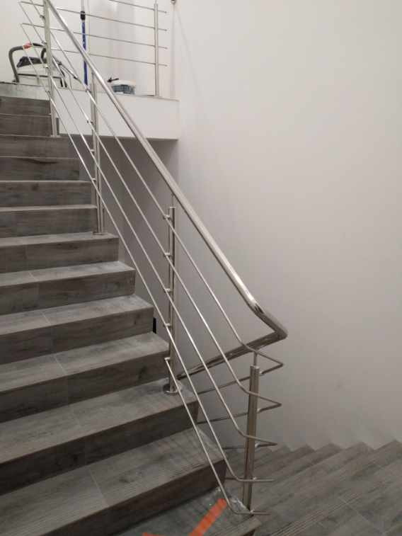 Wewnętrzne schody wyłożone szarymi płytkami imitującymi drewno, zabezpieczone nowoczesną, minimalistyczną poręczą ze stali nierdzewnej z poprzecznymi prętami.
