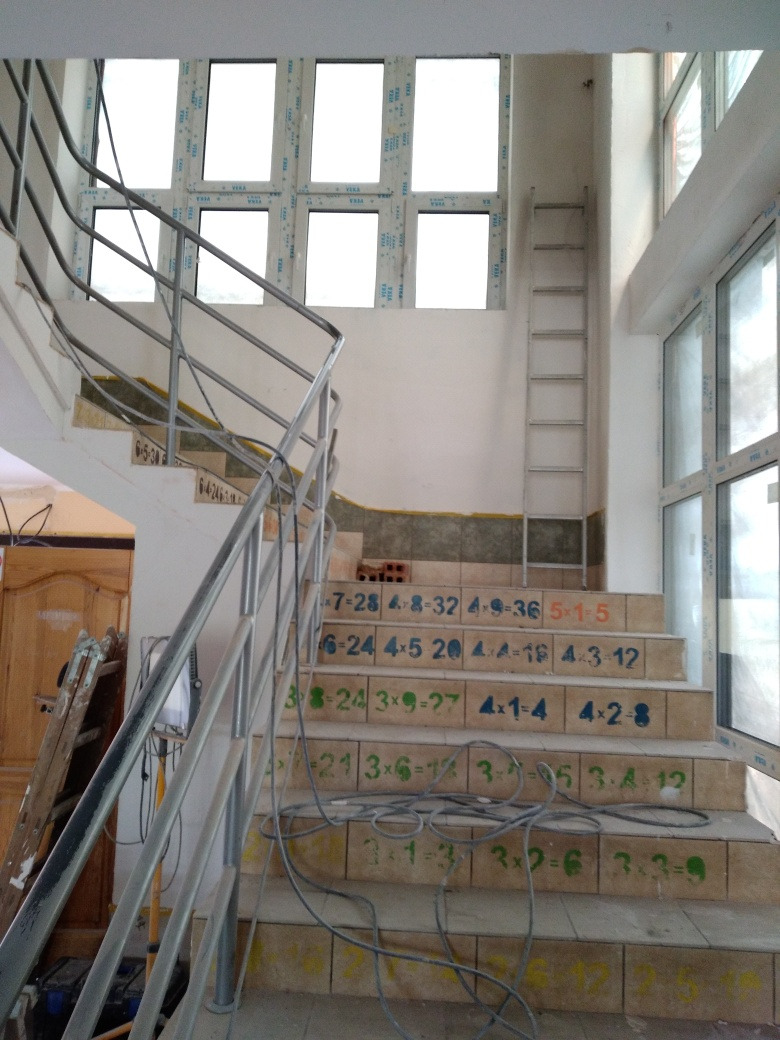 Wnętrze w trakcie remontu z niedokończonymi schodami z ceramicznymi stopniami, na których widnieją równania matematyczne, oraz metalową poręczą. W tle okna i drabina.