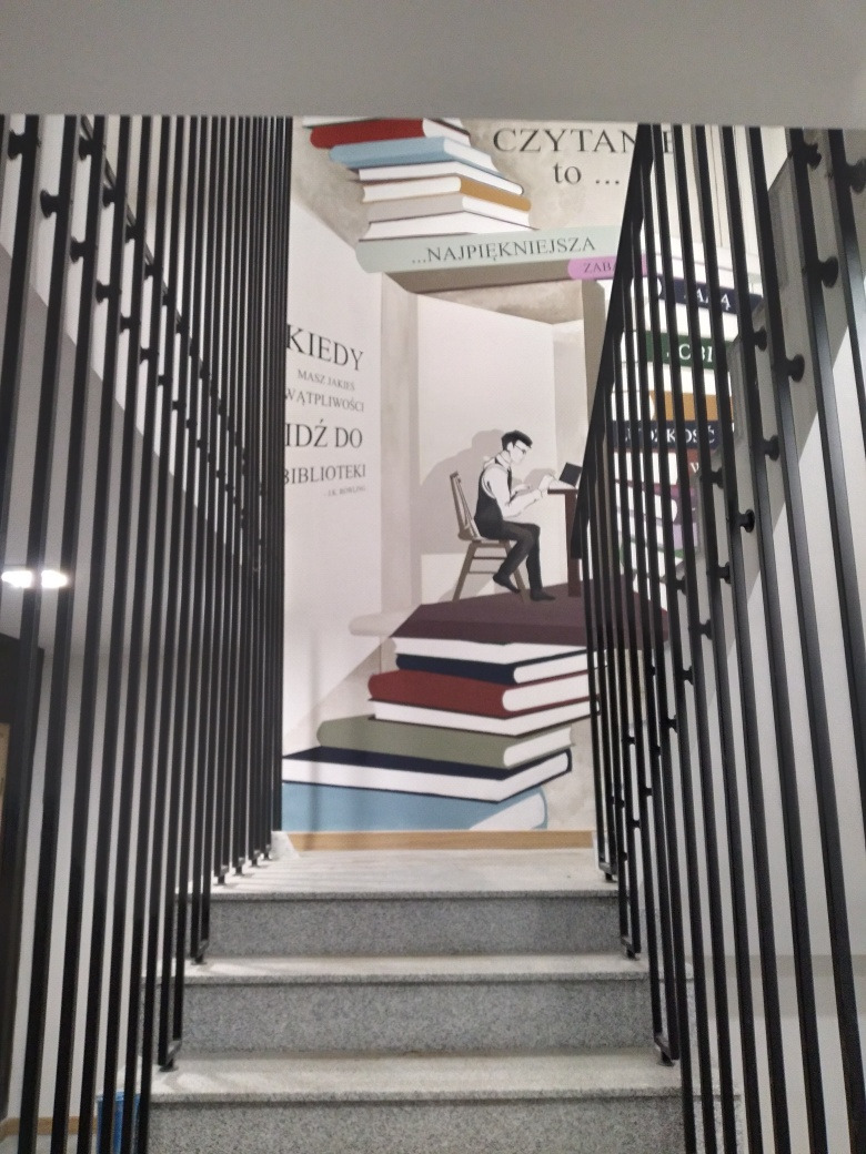 Schody wewnętrzne z szarymi stopniami i czarną metalową balustradą o prostych, pionowych szczeblach, na ścianie dekoracyjny mural z motywem książek i osoby siedzącej przy biurku.