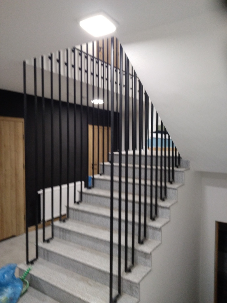 Schody z szarego lastryko z czarną, minimalistyczną balustradą z pionowych prętów, widok z dołu schodów, białe ściany i sufit, oświetlenie sufitowe.