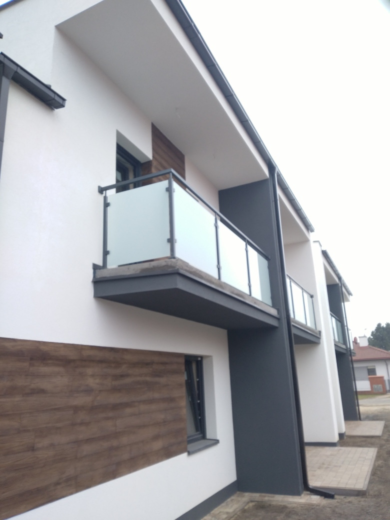 Nowoczesny balkon z balustradą szklaną i ciemnoszarą ramą na tle białej ściany budynku z elementami drewnopodobnymi.