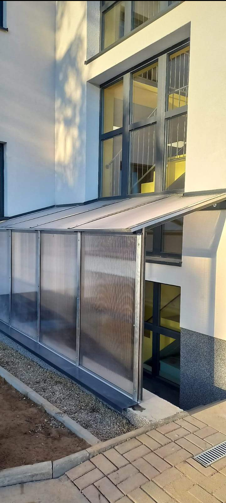 Zadaszenie wejścia do budynku wykonane z paneli z poliwęglanu komorowego w metalowej ramie, widok z zewnątrz, z prawej strony widoczne okna piwniczne z kratami.