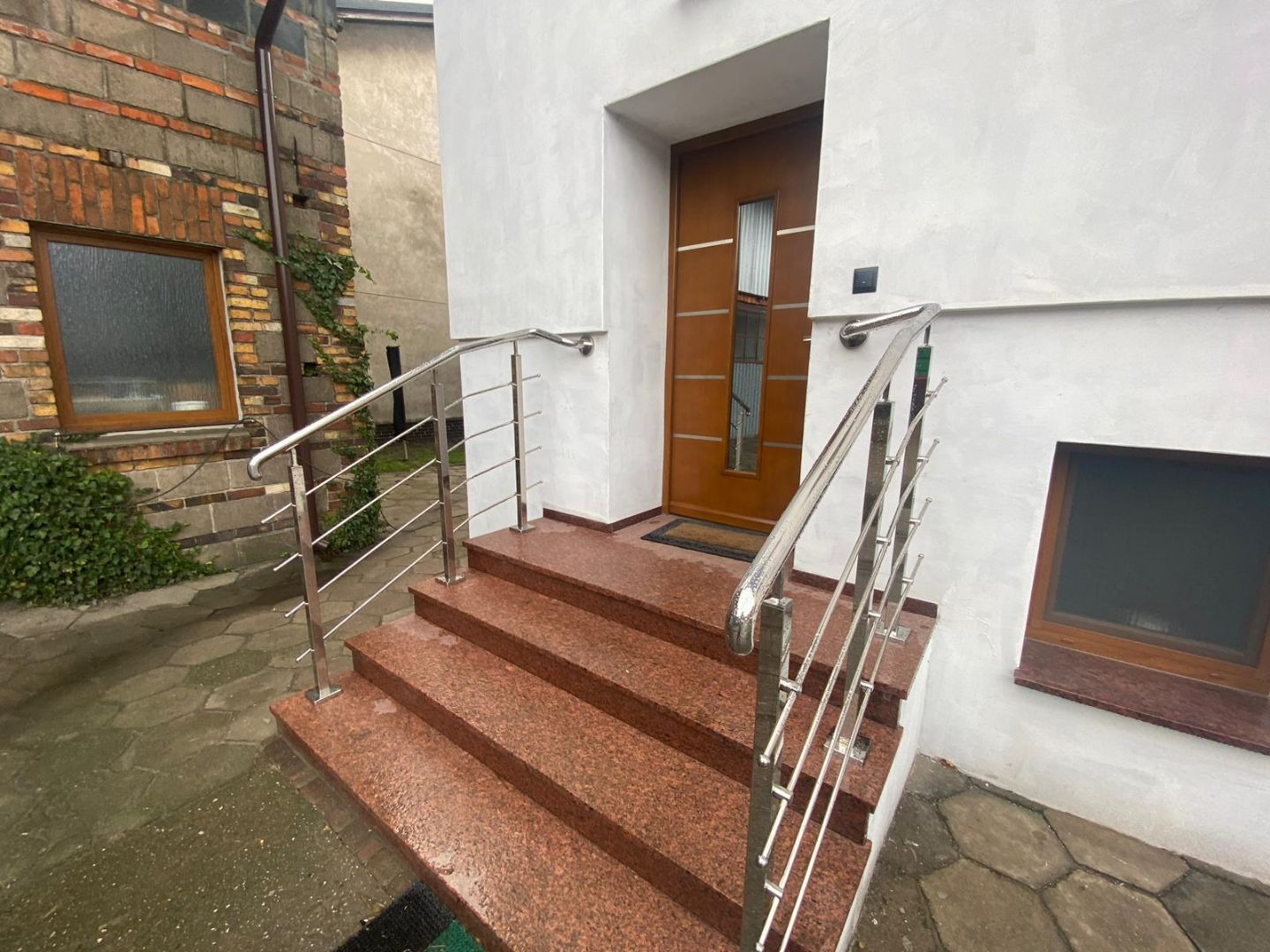 Zewnętrzne schody wejściowe z czerwonego granitu, zabezpieczone nowoczesną, minimalistyczną, metalową barierką z polerowanej stali nierdzewnej, prowadzące do drewnianych drzwi z przeszkleniem.