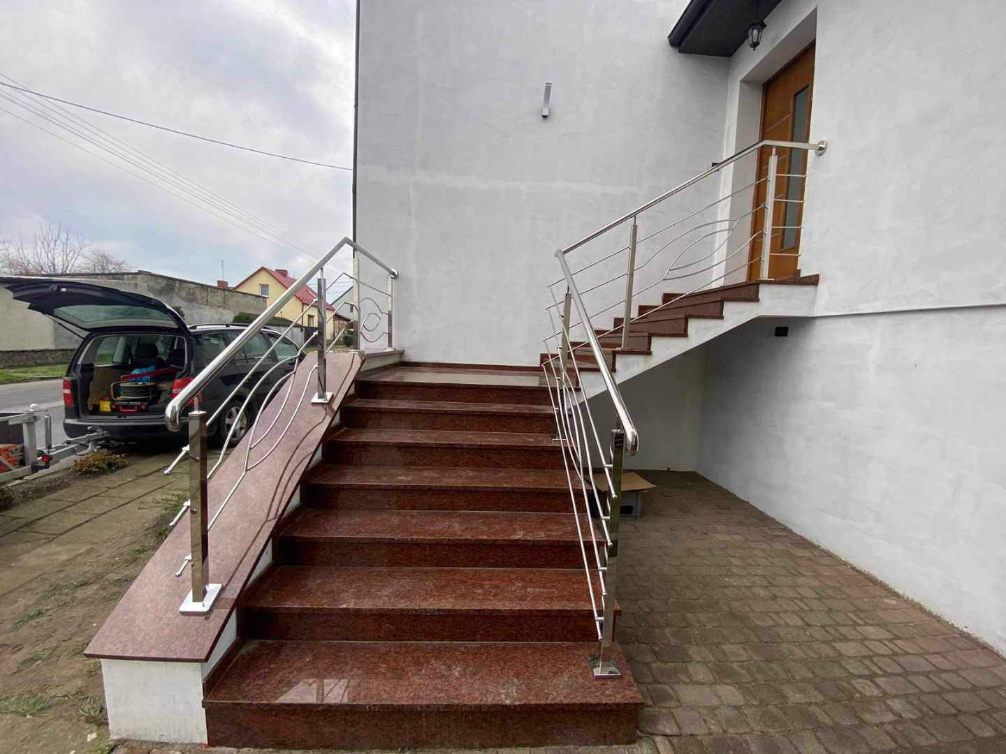 Zewnętrzne schody z czerwonego granitu prowadzące do domu, zabezpieczone nowoczesną balustradą ze stali nierdzewnej z dekoracyjnymi elementami giętymi, na tle zaparkowanego samochodu z otwartym...