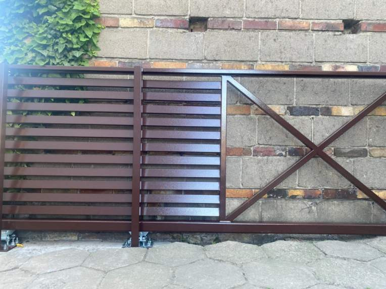 Przesuwna brama w kolorze brązowym z poziomymi panelami i ukośnym wzmocnieniem, zamontowana na szarym bruku na tle ceglanego muru z bluszczem.