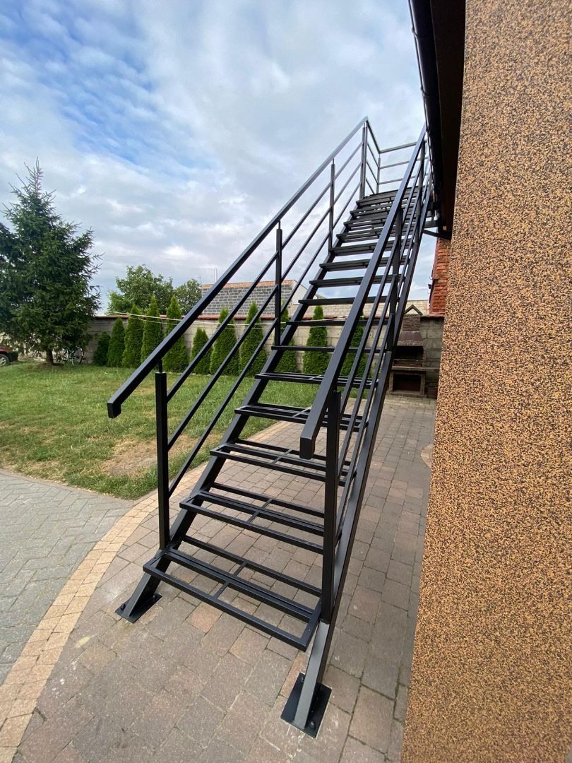 Zewnętrzne schody metalowe w kolorze czarnym, prowadzące do drzwi, z widoczną balustradą i poręczą, na tle budynku o teksturowanej elewacji i zielonego trawnika z tujami.