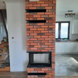 Kominek z cegły klinkierowej z czarną ramką i półkami w nowoczesnym salonie. Widoczne elementy wystroju wnętrza.