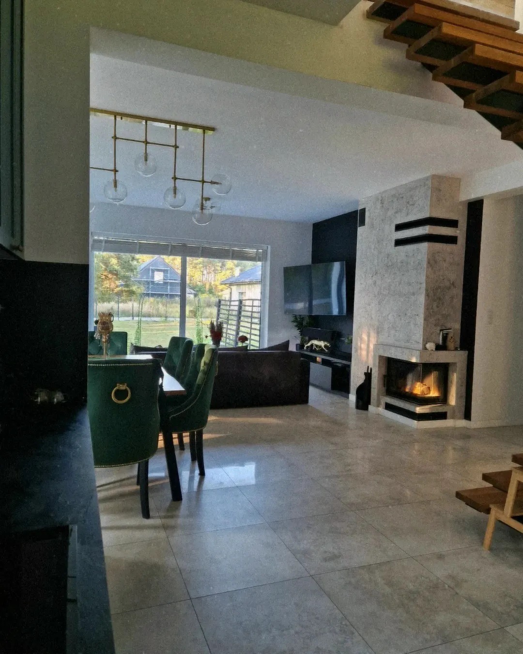 Nowoczesny salon z narożnym paleniskiem obłożonym marmurem, widok z jadalni z zielonymi krzesłami, w tle okno z widokiem na ogród.