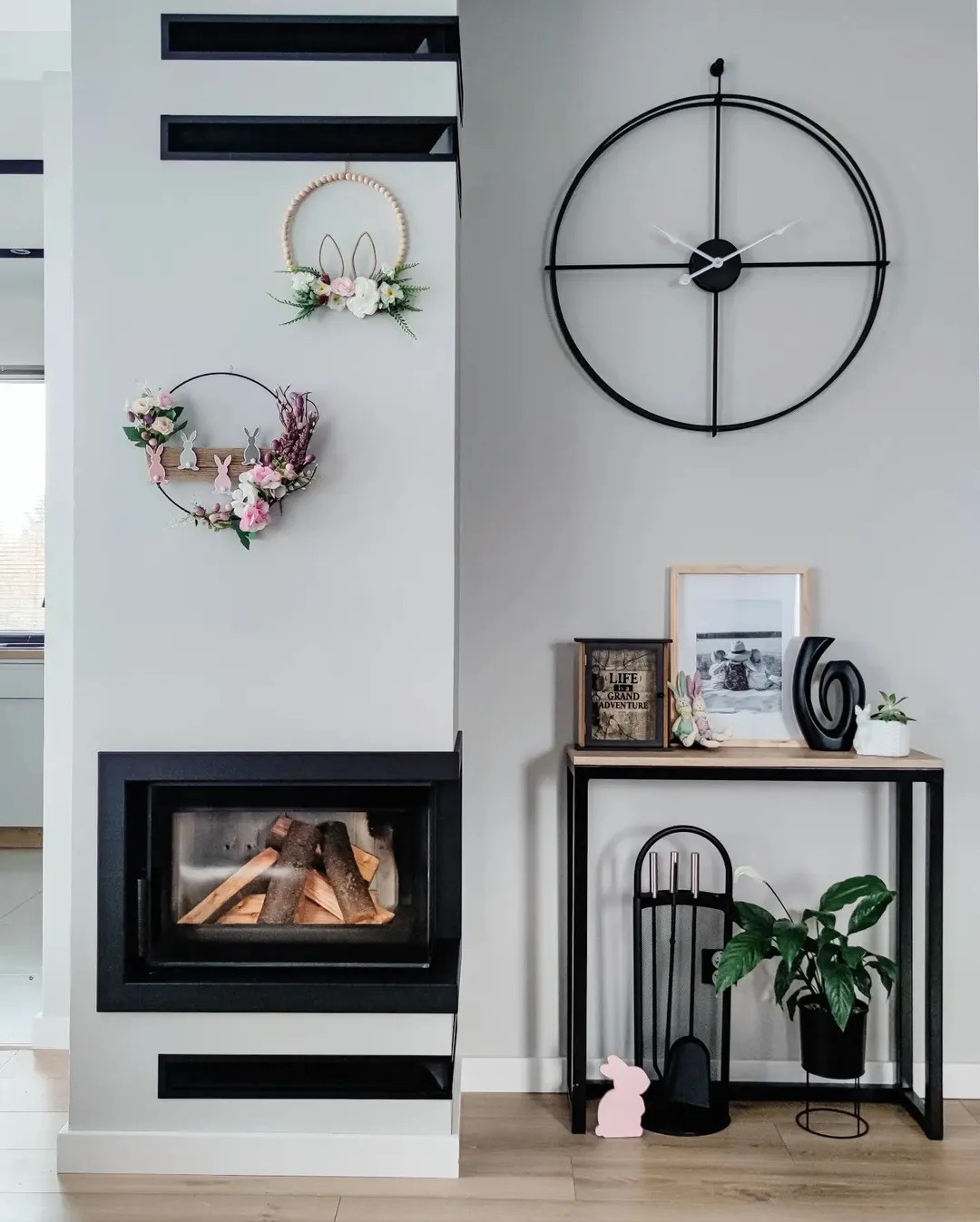 Nowoczesny kominek z czarną obudową, ozdobiony wiosennymi dekoracjami: dwa wieńce z kwiatami i króliczkami na ścianie, duży okrągły zegar oraz konsola z akcesoriami kominkowymi i rośliną doniczkową.
