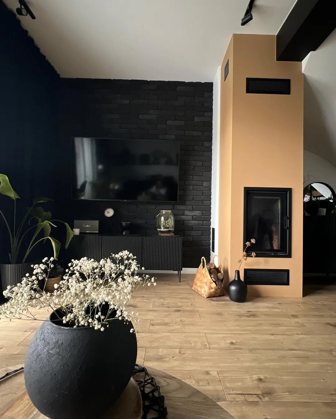 Salon z nowoczesnym, beżowym obudowanym paleniskiem, czarną ceglaną ścianą, telewizorem i czarnymi meblami. Na pierwszym planie czarny wazon z białymi kwiatami.