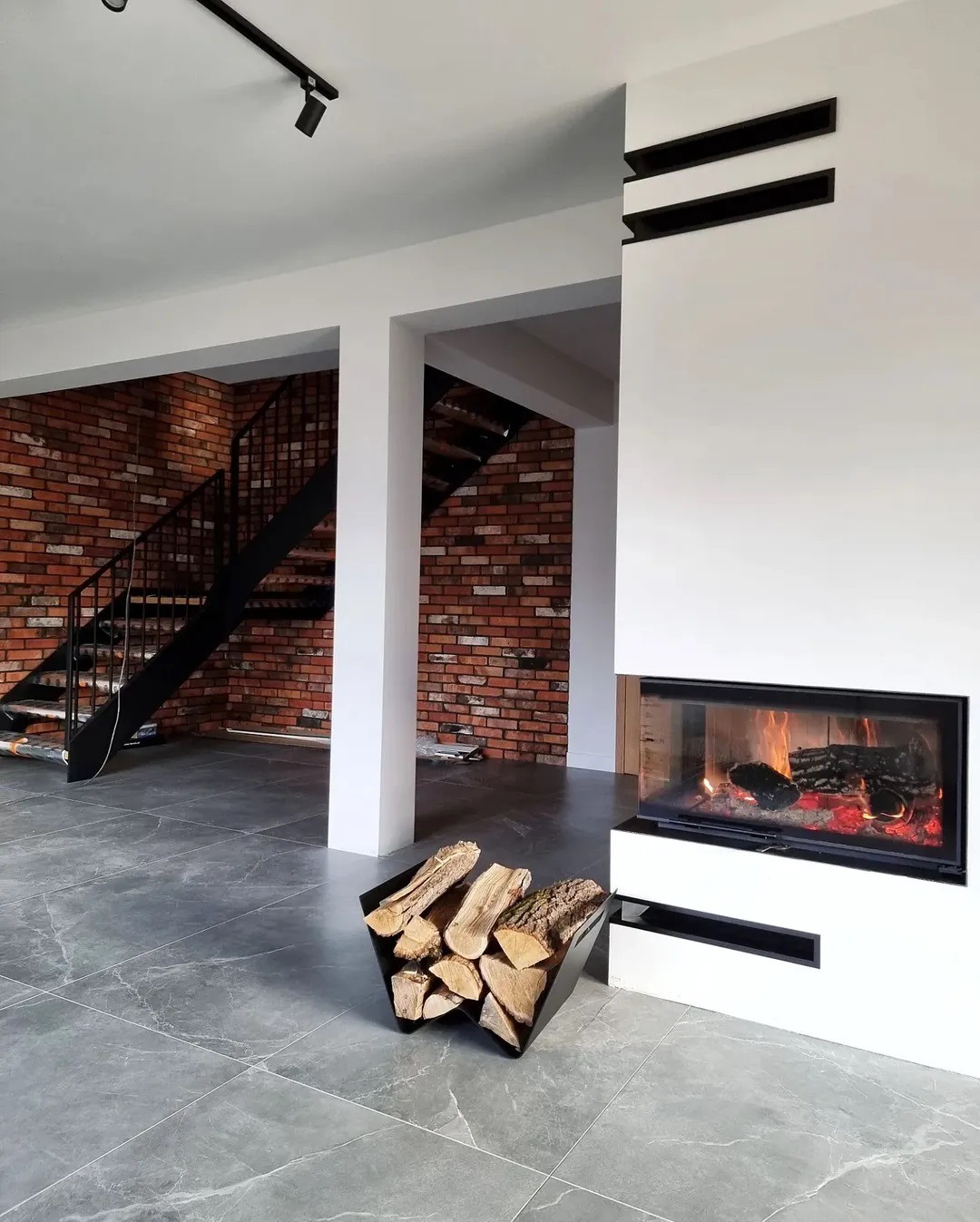 Nowoczesny salon z paleniskiem, widoczne płomienie, kosz z drewnem obok, ceglana ściana i schody w tle.